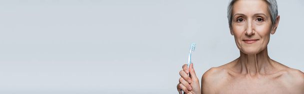 Neşeli orta yaşlı kadın diş fırçasını gri pankartta tek başına tutuyor. - Fotoğraf, Görsel