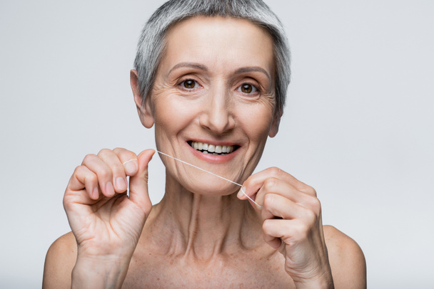 θετική μεσήλικη γυναίκα με γκρίζα μαλλιά που κρατά οδοντικό νήμα απομονωμένο σε γκρι - Φωτογραφία, εικόνα