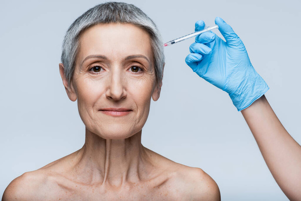 косметолог в латексной перчатке, держащий шприц с инъекцией красоты рядом с улыбающейся женщиной средних лет, изолированной на сером - Фото, изображение