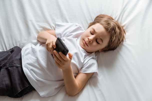 Kleine jongetje ligt op zijn bed en speelt met een smartphone. Close-up portret. Houdt zwarte mobiele telefoon in handen. Concept van online onderwijs. Witte kamer en kleren. Telefoongebruiker. Bespaar kindergezichtsvermogen. - Foto, afbeelding