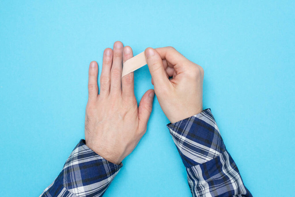 Man in a shirt sticks a medical plaster on his finger. self-help. The concept of first aid for a cut or abrasions on the skin. Blue background - Фото, изображение