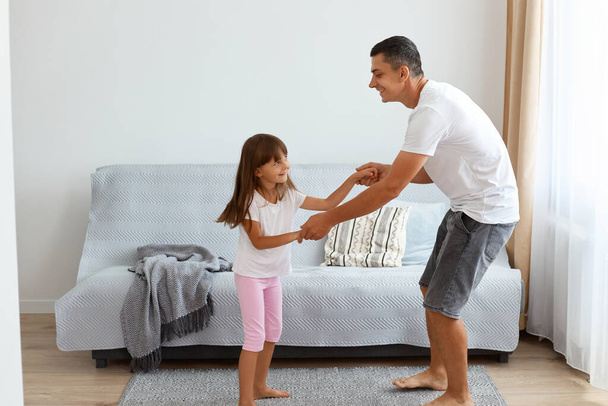 Side view of man wearing white casual style t shirt and jeans short dancing with his daughter and holding her hands, spending happy time together at home. - Фото, изображение