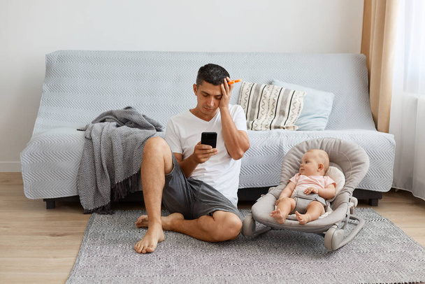 Image of tired exhausted man wearing white casual style T-shirt and jeans short sitting on floor with newborn baby, father didn't sleep whole night, posing closed eyes. - Φωτογραφία, εικόνα