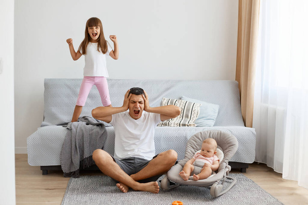 Indoor shot of sad upset depressed man wearing white casual style T-shirt and jeans short sitting on floor with his children, covering ears, being tired of screaming kids. - Zdjęcie, obraz