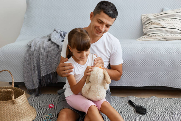 Portrait of smiling happy man wearing white casual style T-shirt and jeans short sitting on floor with daughter, little child holding soft toy, family spending weekend together. - Valokuva, kuva