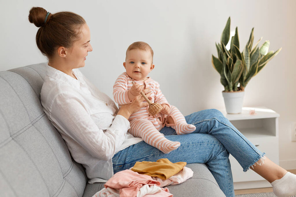 Side view of smiling woman with bun hairstyle wearing white shirt and jeans sitting on sofa with baby daughter, spending time with infant kid, looking smiling at child. - Zdjęcie, obraz