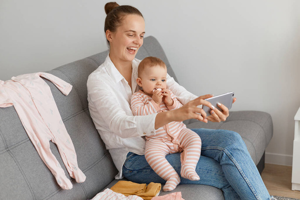 Portrait of attractive happy woman with bun hairstyle wearing white shirt and jeans sitting on sofa with baby daughter and broadcasting livestream, expressing positive emotions. - Foto, Imagen