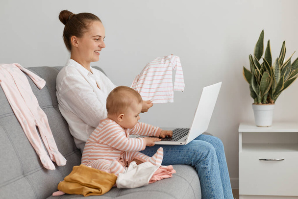 Profile shot of attractive woman with hair bun wearing white shirt and jeans sitting on sofa with baby daughter, holding notebook, showing kid's attire to screen, boasting to her friend her purchase. - Foto, immagini