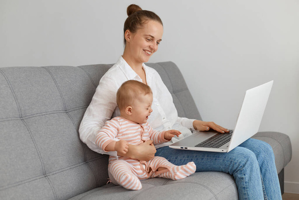 Side view of Caucasian satisfied woman wearing white shirt and jeans sitting on sofa with baby daughter, mother with her kid looking at notebook display with positive facial expression. - Foto, immagini