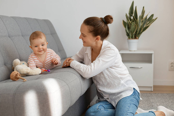 Positive young adult woman with bun hairstyle wearing white shirt and jeans sitting on floor and playing with crawling kid, mother playing with daughter, maternity leave. - Zdjęcie, obraz