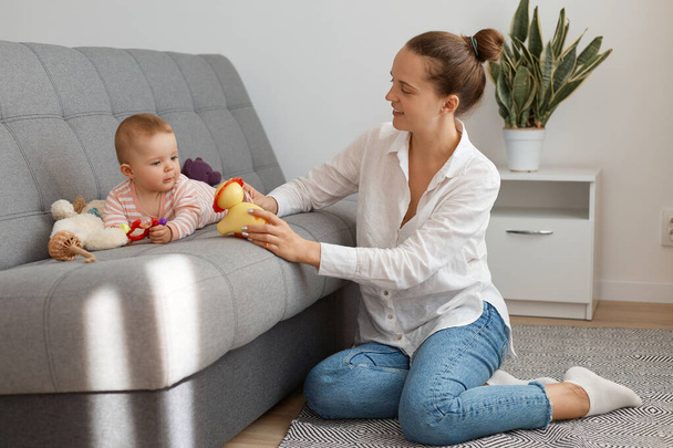Indoor shot of Caucasian woman wearing white shirt and jeans sitting on floor and spending time with infant daughter, mother with bun hairstyle showing toy to child. - Φωτογραφία, εικόνα