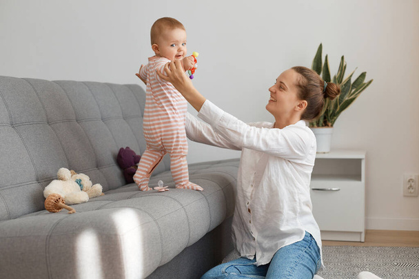 Portrait of delighted woman wearing white shirt and jeans sitting on floor and holding her infant baby wearing striped sleeper, mother playing with her little daughter. - Foto, Bild