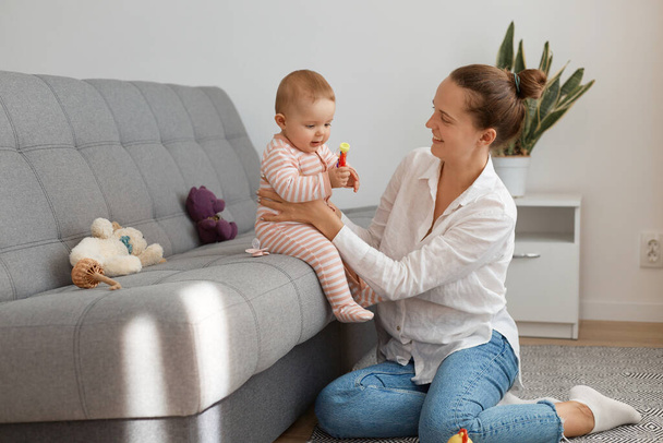 Indoor shot of beautiful woman wearing white shirt and jeans sitting on floor and playing with baby sitting on cough, mother spending time with her daughter, expressing happiness. - Photo, Image