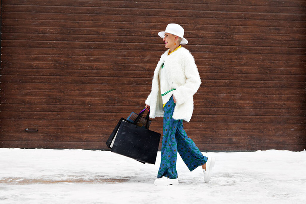 大人の幸せなファッショナブルな女性は、ショッピングバッグ、短い女性の髪、フェルト帽子とスタイリッシュなカジュアル服を着て冬の通りを歩く - 写真・画像