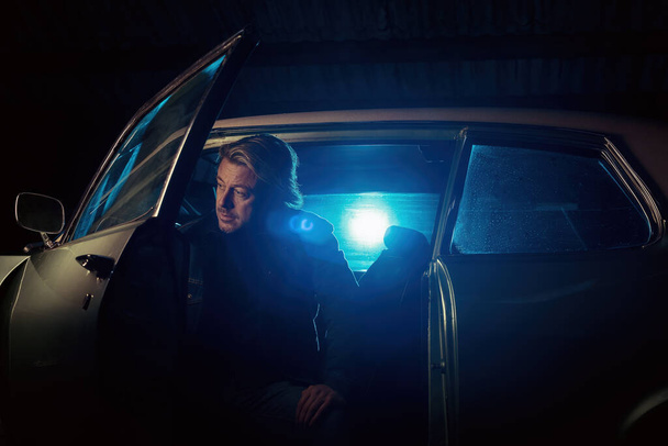 Мужчина с светлыми волосами сидит в классическом американском мускулистом автомобиле с открытой дверью ночью. - Фото, изображение