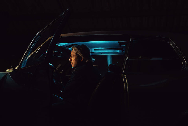 Homem com cabelo loiro senta-se dentro de um carro muscular clássico americano à noite. - Foto, Imagem