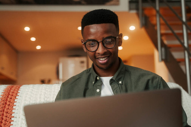 Afrikai amerikai férfi ül a kanapén mosolyogva, miközben gépel a laptop szervez munkát az egyetem  - Fotó, kép
