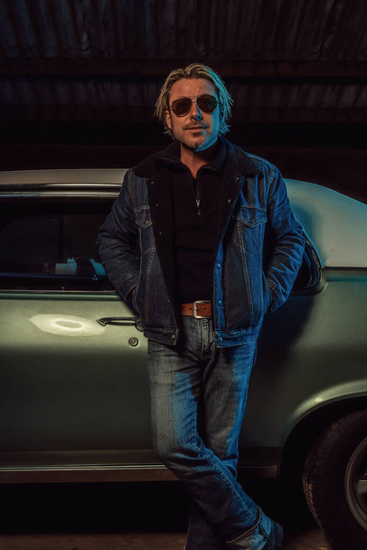 Man with blond hair and sunglasses in a jeans jacket stands by an American classic muscle car. - Foto, Imagem