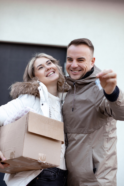 Portrait of smiling couple in casual upper garments stand embracing, holding keys and paperboard box in front of house. - Photo, Image