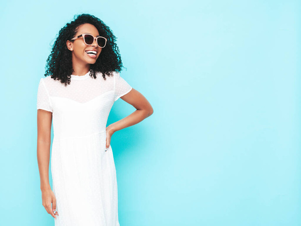 Portrait of beautiful black woman with afro curls hairstyle. Smiling model dressed in white summer dress. Sexy carefree female posing near blue wall in studio. Tanned and cheerful - Zdjęcie, obraz