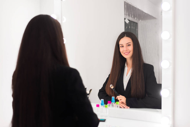 het meisje, een make-up kunstenaar, met lang donker haar, houdt een poeder borstel, kijkt naar zichzelf in de spiegel en glimlacht aan de make-up tafel in een lichte kamer, kantoor - Foto, afbeelding