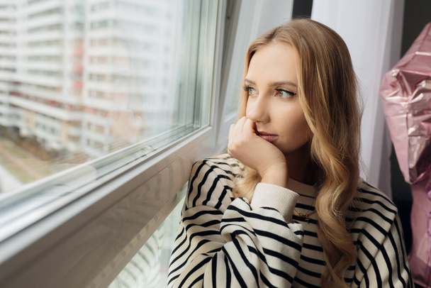 Portrait pensive blond pretty sad depressed woman on windowsill, looking distance. Lonely, tired, isolation. Close up - 写真・画像