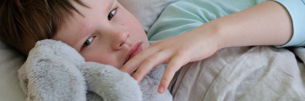 A sad sick boy is lying in bed. Headache and high fever in case of flu or respiratory infection, virus. Epidemic of the disease - Zdjęcie, obraz