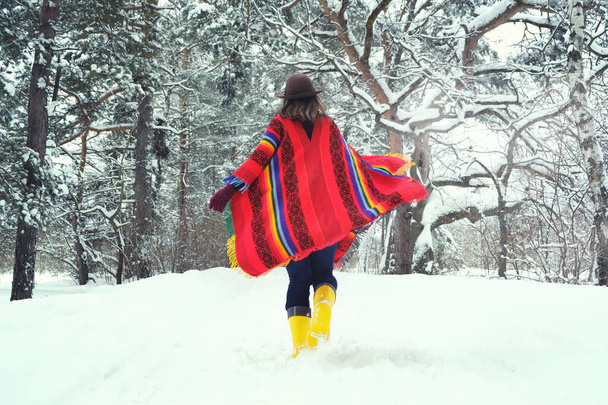 View from behind woman in colorful wool ethnic poncho and hat running in snowy winter forest. Boho style fashion. Enjoing nature at cold season.  - Photo, image