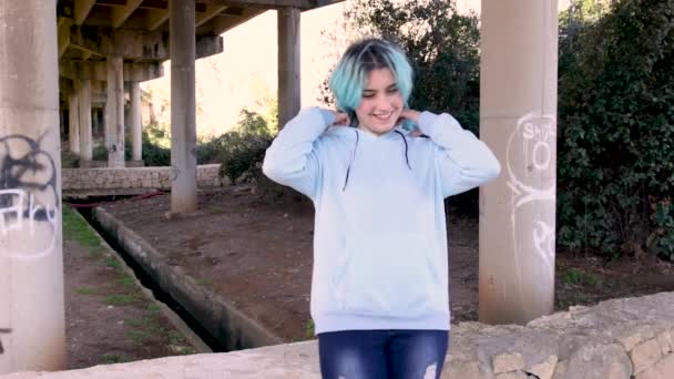 Adolescente sonriente que usa sudadera con capucha y se mantiene contra pilares del puente. Chica adolescente de pelo azul se queda al aire libre contra los pilares del puente. Maqueta de ropa - Imágenes, Vídeo