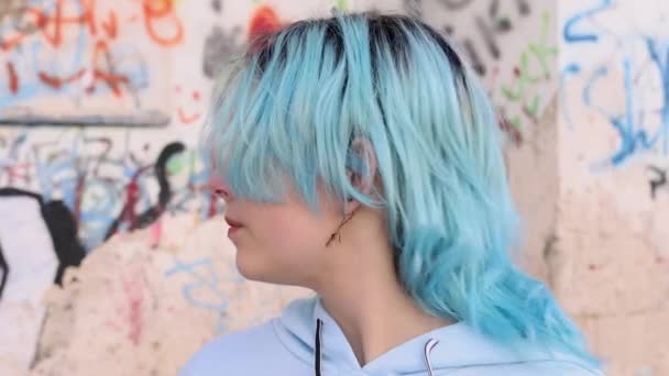 Σοβαρή Teenager σε γαλάζιο oversized hoodie στρίψτε αριστερά. Μπλε μαλλιά έφηβος κορίτσι που διαμένουν σε εξωτερικούς χώρους κατά γκράφιτι στον τοίχο και κοιτάζοντας στην κάμερα. Πυροβολισμός στο κεφάλι - Πλάνα, βίντεο