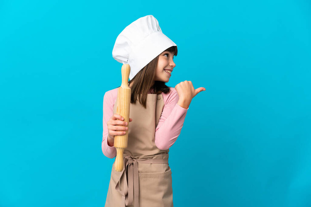 Little girl holding a rolling pin isolated on blue background pointing to the side to present a product - Photo, Image