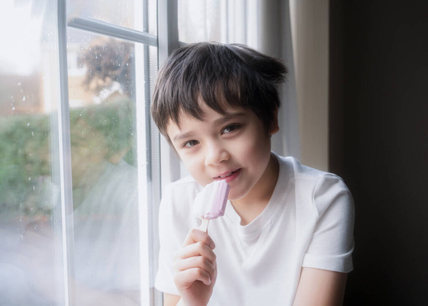 Egy boldog gyerek jégkrémet eszik, portré egy jóképű fiatal fiúról, aki az ablak mellett ül és frissítőt iszik, egy mosolygó arcú gyerek, aki otthon pihen. A srác a kamerába néz. - Fotó, kép