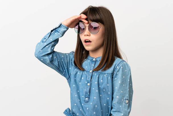 Κοριτσάκι με γυαλιά ηλίου απομονωμένο σε λευκό φόντο κάνει χειρονομία έκπληξη, ενώ κοιτάζοντας προς τα πλάγια - Φωτογραφία, εικόνα