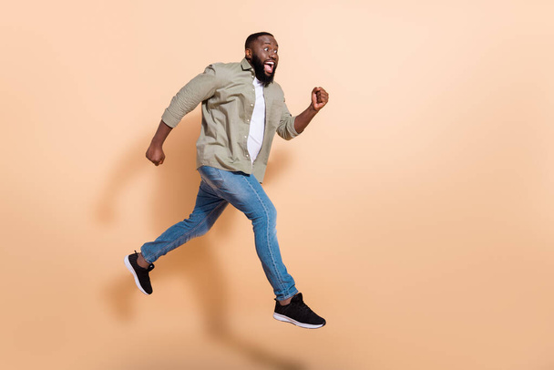 Full size profiel foto van onder de indruk millennial brunet jongen springen schreeuwen dragen shirt jeans sneakers geïsoleerd op beige achtergrond - Foto, afbeelding