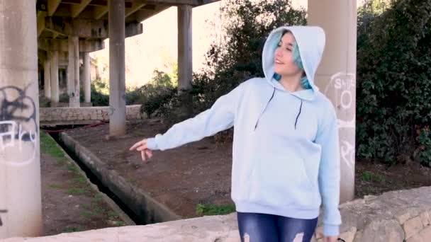 Teenager in light blue oversize hoodie and jeans Smiling and dancing. Blue haired teen girl stays outdoors against bridge pillars. Clothing mockup - Footage, Video