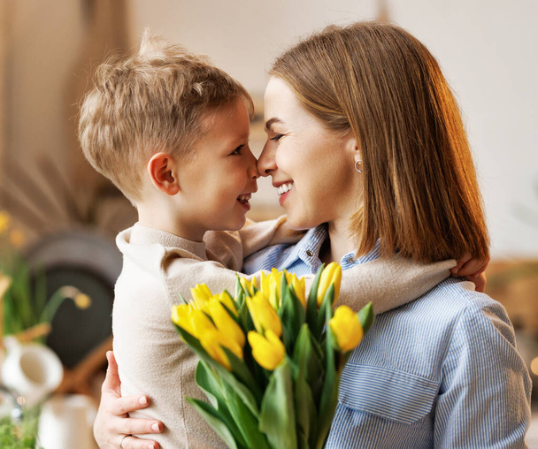 Junge Mutter mit Blumenstrauß umarmt Sohn, während sie Glückwünsche zum Muttertag entgegennimmt - Foto, Bild