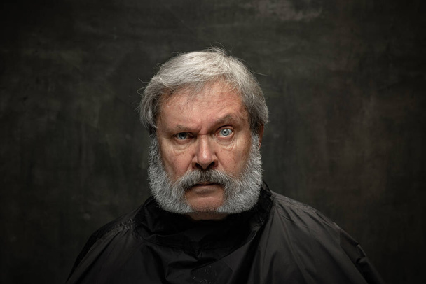 Dramatic portrait of emotional senior bearded man getting beard grooming isolated on dark vintage background. Concept of emotions, fashion, beauty, self-reinvention - Fotó, kép