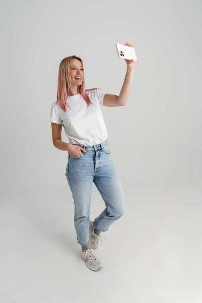 Happy young woman in casual wear standing taking selfie holding mobile phone over gray background, full length - Foto, Imagen