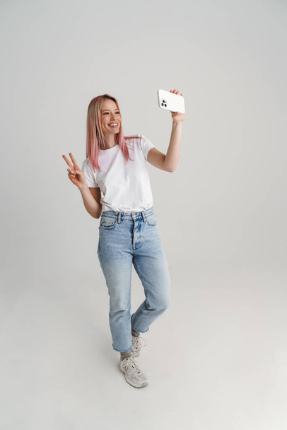 Mujer joven feliz en ropa casual de pie tomando selfie celebración de teléfono móvil sobre fondo gris, longitud completa - Foto, imagen