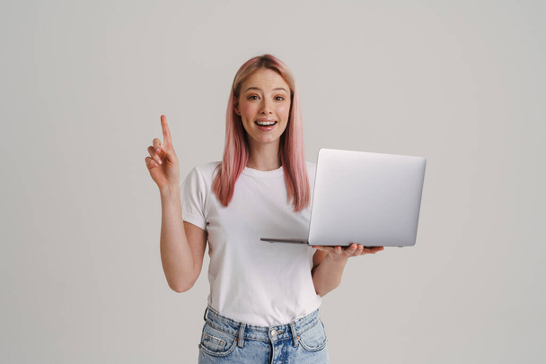 Young woman smiling while holding laptop and pointing finger upward isolated over white background - Foto, imagen