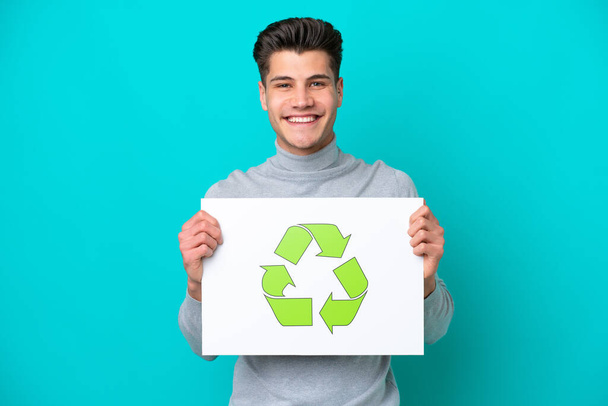 Jeune homme caucasien beau isolé sur bakcground bleu tenant une pancarte avec icône de recyclage avec expression heureuse - Photo, image