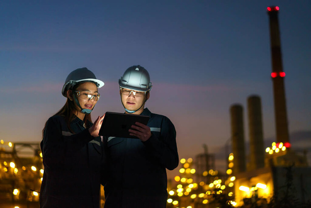 Ingenieros asiáticos, hombre y mujer están comprobando el mantenimiento de la fábrica de refinería de petróleo por la noche a través de tabletas digitales - Foto, Imagen