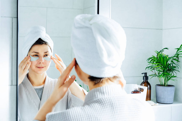 Woman in a bathrobe with a towel on her head applying patches under eye area, looking at mirror, enjoying daily antiwrinkle antiage routine in bathroom after morning shower. Home Beauty self-care - Zdjęcie, obraz