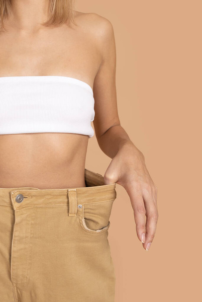 Close-up of slim Caucasian woman wearing oversize pants and white bra underwear showing slender waist on skin colored background. Weight loss. Keeping a healthy diet, exercising - Photo, Image