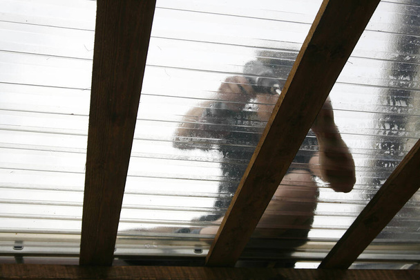 屋外のホームテラスにクリアPVC屋根シートを取り付けるために電動工具を使用して作業する男. - 写真・画像