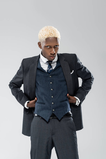 blonde african american man looking at camera and posing with hands on waist isolated on grey  - Fotografie, Obrázek