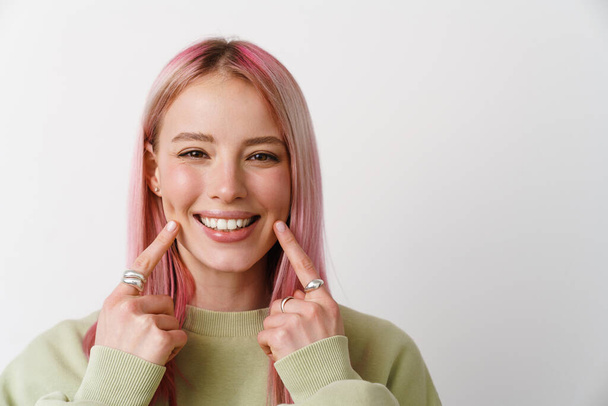 Young smiling woman with pink hair pointing finger at her cheek isolated over white background - 写真・画像