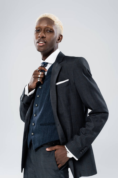 blonde african american man in formal wear adjusting tie and posing with hand in pocket isolated on grey  - Fotografie, Obrázek