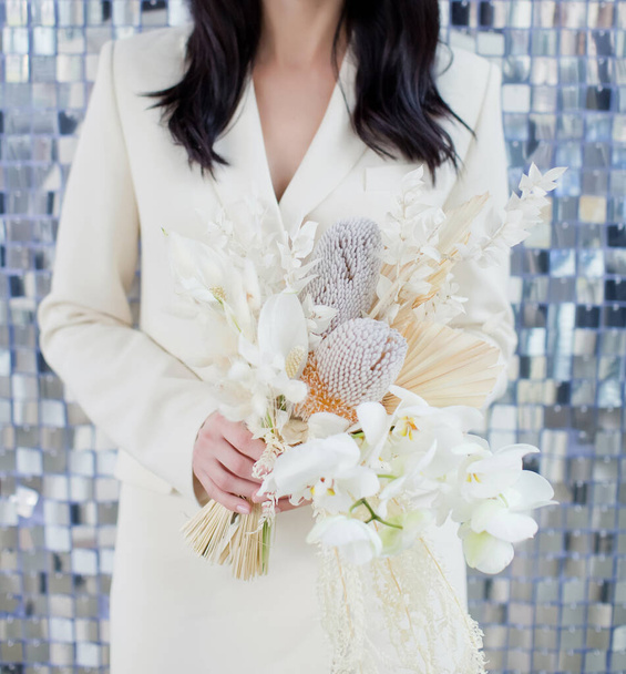Bride in beige suit holds wedding boho bouquet with orchids, banksia and dry flowers on a silver sequin texture background - Photo, Image
