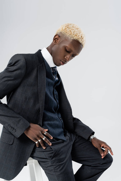 blonde african american man in suit leaning on chair isolated on grey  - Fotografie, Obrázek